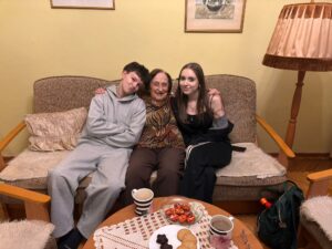 Trzy osoby siedzą obok siebie na kanapie w domowym salonie. Po obu stronach stoją drewniane fotele, a obok znajduje się wysoka lampa z abażurem. Na stoliku przed kanapą leży serwetka i ustawione są kubki, talerzyk z ciastkami oraz miseczka ze słodyczami. W tle wiszą dwa obrazy na jasnej ścianie.