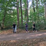 Trzy osoby idą leśną ścieżką, zbierając śmieci wśród drzew. Jedna z nich trzyma duży, przezroczysty worek. W tle widać gęste, zielone drzewa i krzewy