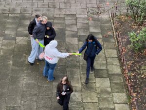 Na zdjęciu widać grupę kilku osób stojących i przechodzących po chodniku z betonowych płyt. Jedna osoba w jasnej bluzie wyciąga w stronę przechodnia długi, jaskrawy przedmiot. Obok stoją dwie inne osoby, jedna trzyma podobny przedmiot. Na pierwszym planie idzie kolejna osoba z długimi włosami, trzymając coś w ręku. W tle znajdują się niskie krzewy oraz jesienne liście leżące na ziemi.