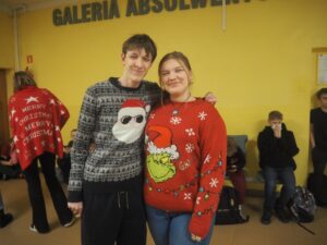 Dwie osoby stoją w szkolnym korytarzu na tle żółtej ściany z napisem ‘Galeria Absolwentów’. Mają na sobie świąteczne swetry: jeden z motywem Mikołaja, drugi z postacią Grincha. W tle widać kilka innych osób siedzących na ławkach oraz osobę przechodzącą w czerwonej pelerynie z napisem ‘Merry Christmas