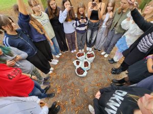 Grupa młodych osób stoi w kręgu na leśnej ścieżce, patrząc na kilka dużych worków wypełnionych kasztanami ułożonych na ziemi. Wokół widać opadłe liście, a część uczestników trzyma w rękach telefony.