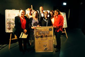 Grupa osób stoi w sali wystawowej przy dużym plakacie zatytułowanym „Wczoraj”, a w tle znajdują się dodatkowe plansze prezentacyjne na sztalugach.