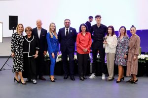 Grupa osób stoi razem na tle jasnego ekranu w sali wydarzeniowej, ustawiona w jednym rzędzie przed dekoracją z białych kwiatów. Jedna z osób trzyma statuetkę lub nagrodę.