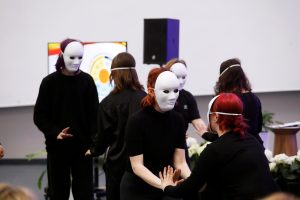 Grupa osób ubranych na czarno wykonuje scenę teatralną lub ruchową, stojąc w parach i trzymając się za ręce w jasnym pomieszczeniu wydarzeniowym. W tle widać ekran z kolorową grafiką oraz głośnik.