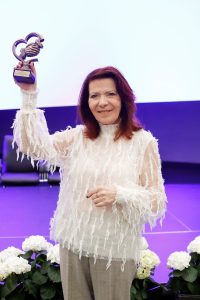 Osoba stoi na scenie i unosi w górę statuetkę w kształcie serca, ubrana w jasny, fakturowany top. Na pierwszym planie znajdują się białe kwiaty, a w tle widać jasne oświetlenie sceny.