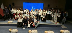 Zdjęcie przedstawia dużą grupę młodzieży i dorosłych pozujących do wspólnej fotografii na scenie w nowoczesnej sali konferencyjnej. Wydarzenie ma charakter edukacyjny, związany z kursem pierwszej pomocy.