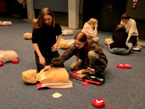Zdjęcie przedstawia młodzież podczas praktycznych ćwiczeń z zakresu pierwszej pomocy, odbywających się w dużej sali o niebieskiej wykładzinie.