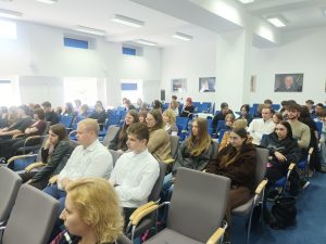 Zdjęcie przedstawia szeroki plan na widownię wypełniającą salę konferencyjną. Kadr wykonany jest z boku, ukazując rzędy siedzących osób.