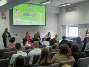 Zdjęcie dokumentuje debatę odbywającą się w nowoczesnej sali wykładowej. Kadr obejmuje panelistów siedzących przodem do widowni oraz plecy uczestników zgromadzonych w rzędach krzeseł.