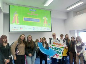 Zdjęcie przedstawia grupę uśmiechniętych młodych ludzi pozujących w sali wykładowej na tle dużego ekranu projekcyjnego.