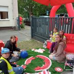 Grupa dzieci siedzi i klęczy na sztucznej trawie pod czerwonym namiotem podczas wydarzenia plenerowego. Uczestnicy układają duże puzzle z napisem związanym z krwiodawstwem. Wokół widać elementy stoiska, w tym czerwone dekoracje i materiały promocyjne. W tle znajdują się ogrodzenie, zaparkowane samochody oraz dmuchana instalacja reklamowa z symbolem krzyża.