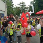 Na zewnątrz odbywa się wydarzenie promocyjne, podczas którego grupa dzieci w odblaskowych kamizelkach stoi i porusza się wokół stanowiska związanego z akcją krwiodawstwa. Dzieci trzymają czerwone balony z symbolem krzyża oraz przekąski, a wśród nich znajduje się osoba w dużym kostiumie kropli krwi. W tle widoczny jest czerwony namiot z napisem „Twoja krew – darem życia”, dmuchana atrakcja oraz budynki i drzewa.
